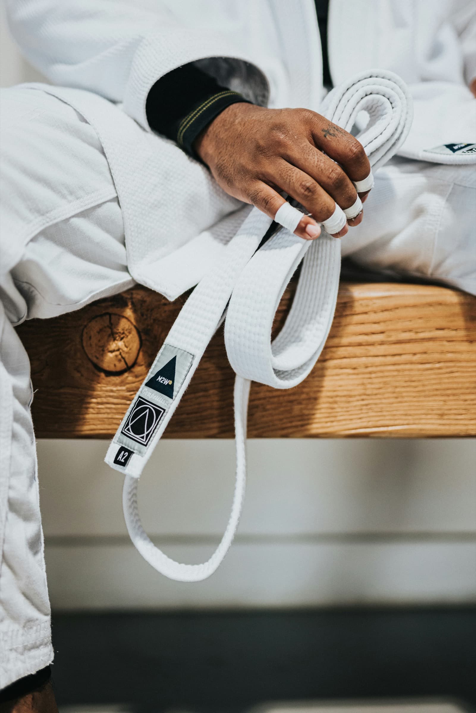 Martial artist in a white gi holding a belt, seated on a wooden bench
